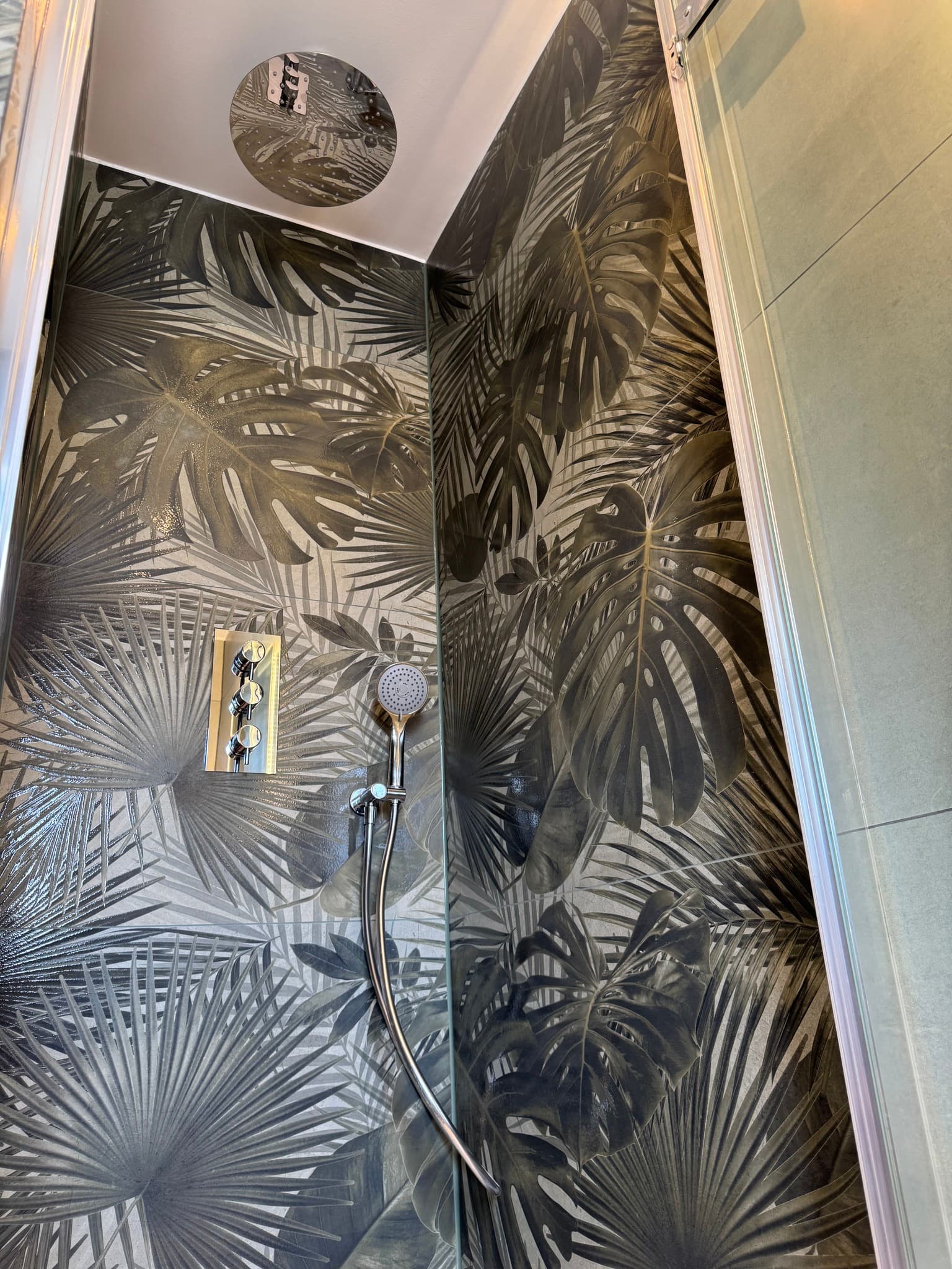 Shower enclosure fully tiled in dark tropical leaf-print ceramic tiles with chrome thermostatic shower valve, rail, and ceiling-mounted rain head