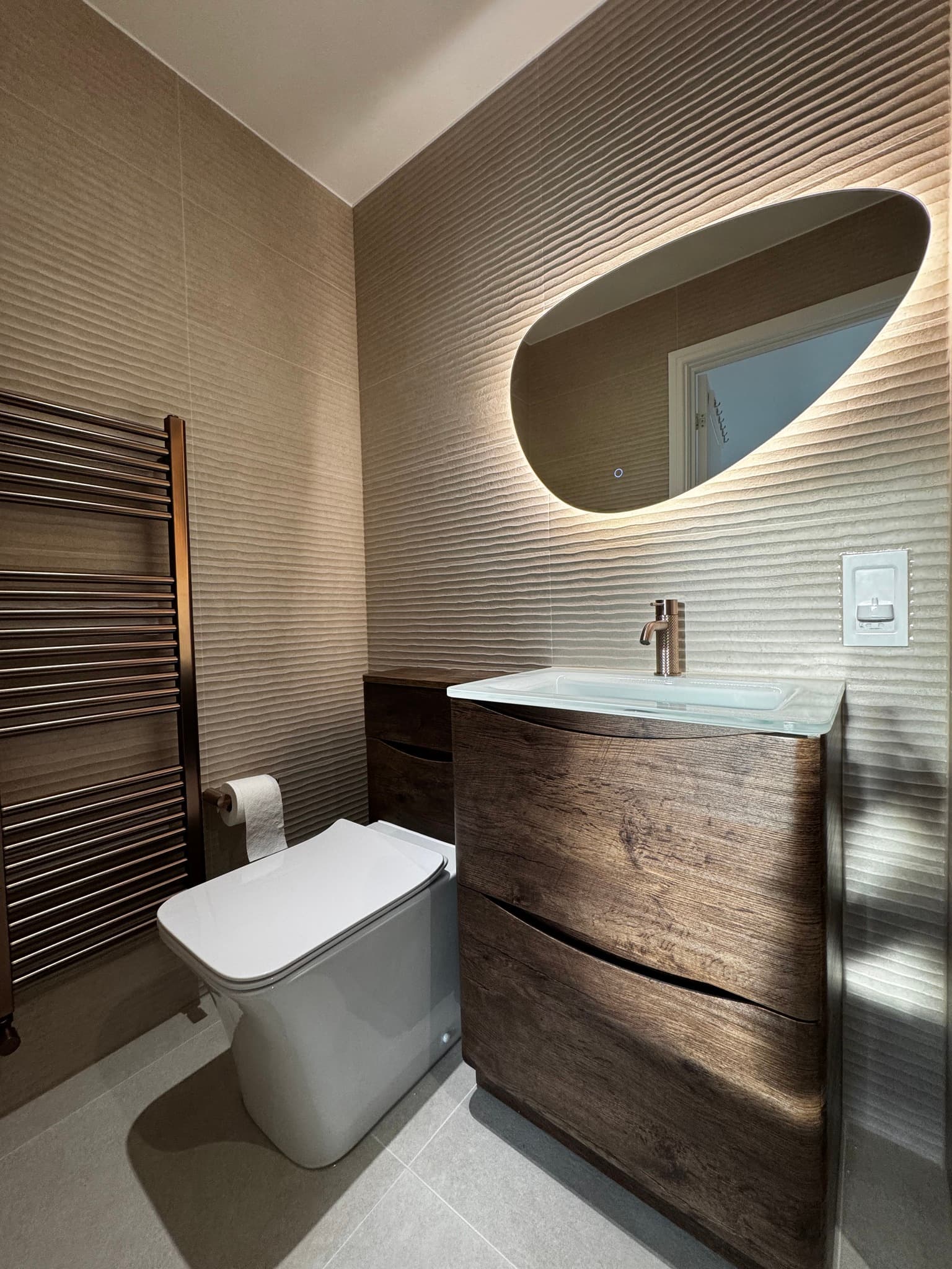 Warm bathroom detail showing sand wave-texture tiles and bronze heated towel rail — ABC Tiling craftsmanship