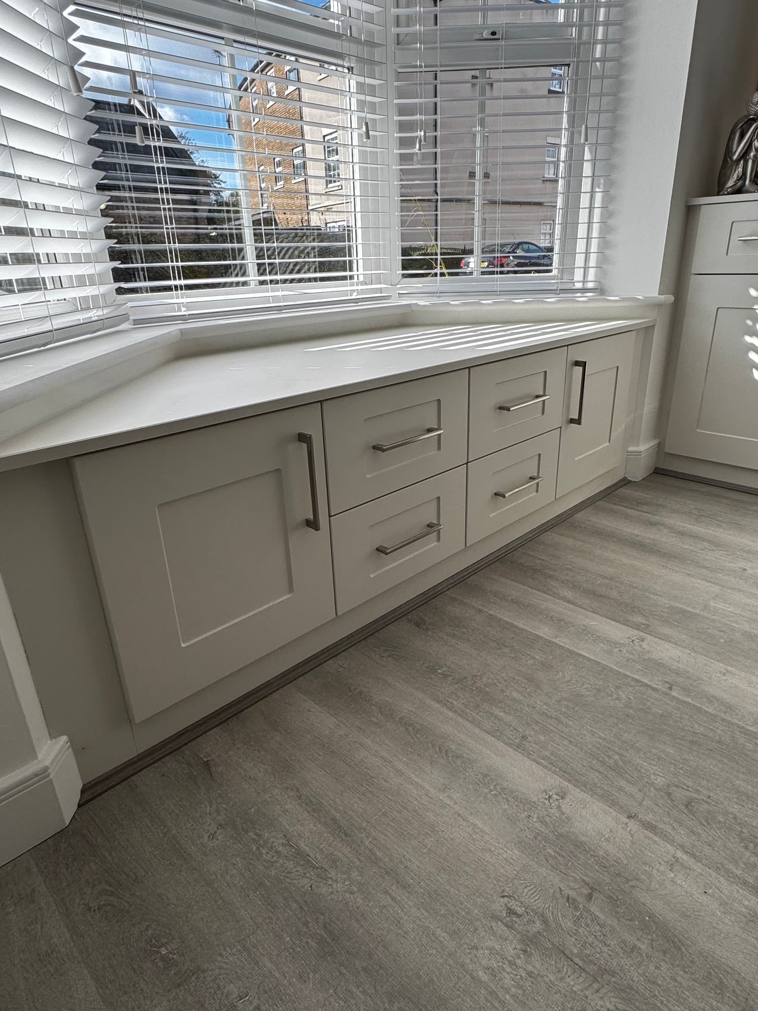 Kitchen bay window area with cream shaker base units, white quartz worktop, and grey wood-effect flooring