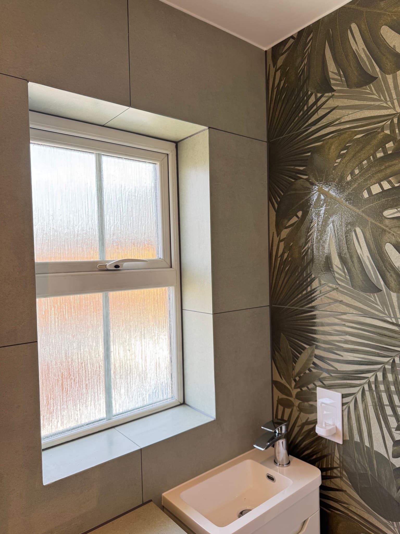 Bathroom window reveal tiled in stone-effect tiles beside a bold tropical leaf-print ceramic feature wall